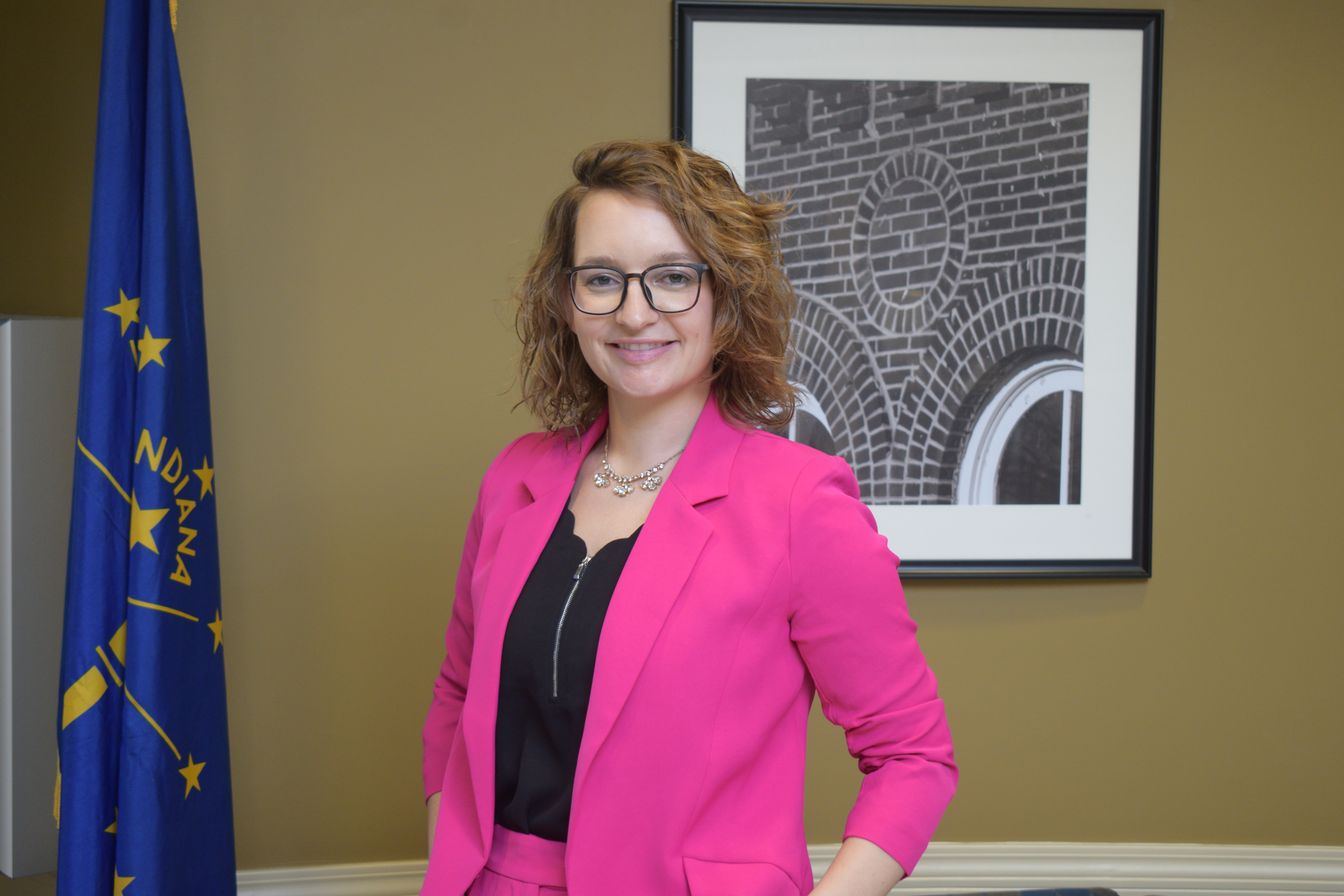 Sarah McLin wearing a pink suit, black blouse and glasses stands next to a State of Indiana flag with a framed piece of art behind her.