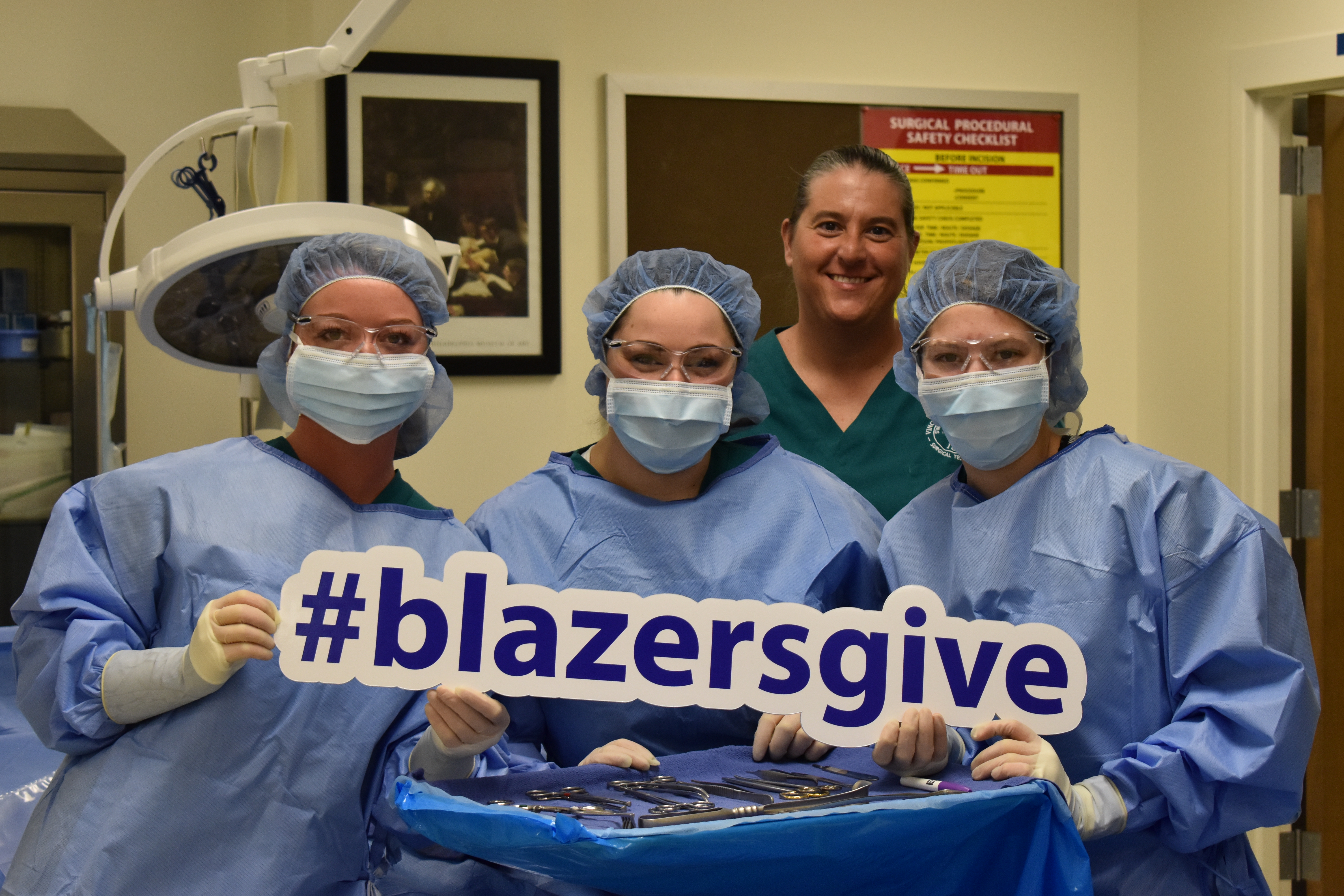 Three female Surgical Technology students and a female Surgical Technology professor pose with a #BlazersGive sign wearing surgical gowns, scrubs, masks and protective eyewear
